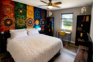 A bedroom with a bed, a desk, a chair, a bookshelf, a ceiling fan, and a window with a view of greenery.