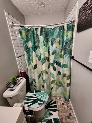Un bagno con tenda da doccia decorata con un motivo a foglia, un water, una piccola pianta su uno scaffale e un pavimento piastrellato.