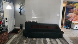 A modern living room with a wooden floor, a black sofa, a wall-mounted art piece, and a door with a frosted glass window. - The Vibe House - 3BR Island Retreat near Savannah & Tybee (Savannah)