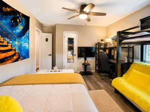 A bedroom with a bed, a desk, a chair, a bunk bed, a ceiling fan, and a painting on the wall.