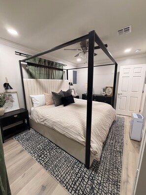 Un dormitorio con cama con dosel, una mesita de noche con lámpara, una mesita auxiliar, una alfombra estampada y una puerta blanca.