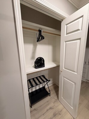 Un pequeño espacio de almacenamiento cerrado con una puerta blanca, una estantería con un ventilador negro y un mueble negro con estantes.