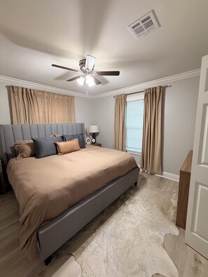 Une chambre à coucher avec un lit, un ventilateur de plafond, une table de chevet, une lampe et une fenêtre avec des rideaux.