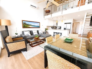 Ein modernes Wohnzimmer mit einer Couch, einem Couchtisch und Korbstühlen. Im Hintergrund ist eine Küche mit einem Essbereich erkennbar.