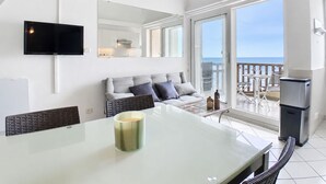 Una sala de estar moderna con un sofá, un televisor de pantalla plana, una mesa de vidrio con una vela, y un balcón con vista al mar.