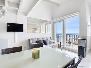 Ein modernes Wohnzimmer mit Sofa, Flachbildfernseher, Glastisch mit Kerze und Balkon mit Meerblick.