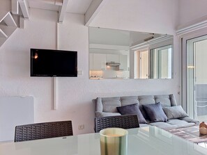 A modern living room with a flat-screen TV, a sofa, a glass coffee table, and a kitchen in the background. - Ortal Océan by Interhome (Lacanau)
