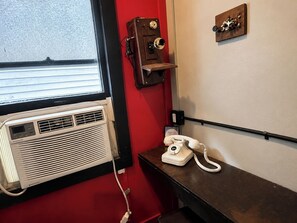 Huoneessa on ikkuna, seinään kiinnitetty puhelin, lämmitin ja pieni hylly, jonka päällä on koriste-esine.