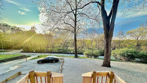 Une terrasse en bois mène à un chemin de gravier, et on aperçoit une voiture garée au loin.