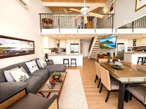 A spacious living area with a gray sectional sofa, a coffee table, and a dining table with woven chairs. A staircase connects the living area to the upper floor. - Napili Point 3 Waterfront Villas, Sleeps 16 | Car Incl. w/ 6 nights | ML-4083 by KBM (Lahaina)