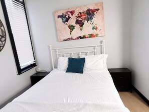 A neatly made bed with white bedding and a blue pillow, a world map artwork on the wall, a window with blinds, and a small bedside table.