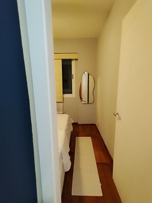Un couloir étroit mène à une chambre qui contient un lit, une fenêtre avec des stores, un miroir et une porte.