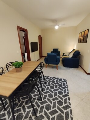 Ein Essbereich mit Holztisch und -stühlen, einem Topf mit Pflanze, einem gemusterten Teppich, einem blauen Sofa und einem Deckenventilator.
