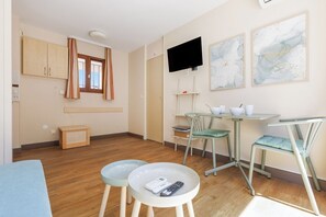 Ein Zimmer mit Holzboden, einem an der Wand montierten Fernseher, einem kleinen Tisch mit Stühlen und einem Fenster mit Vorhängen.