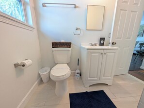 A bathroom with a toilet, a sink, a mirror, and a trash bin. - The Beach Shack - 1BR Neptune Getaway (Neptune Beach)