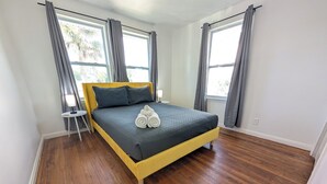 A bedroom with a bed, two pillows, a bedside table with a lamp, and a window with curtains. - The Beach Shack | 1BR Getaway (Neptune Beach)
