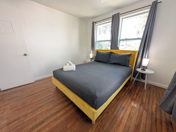 A bedroom with a bed, a nightstand, a lamp, and a window with curtains. - The Beach Shack - 1BR Neptune Getaway (Neptune Beach)
