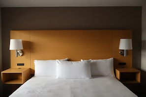 A hotel room with a wooden headboard, two bedside tables, a bed with white linens, and wall-mounted lamps.