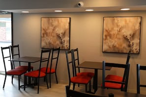 Un espace repas avec des tables et des chaises, deux Ćuvres dâart encadrĂ©es accrochĂ©es au mur et une fenĂȘtre laissant entrer la lumiĂšre naturelle.