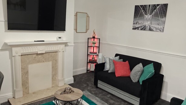 Een woonkamer met een open haard, een zwarte bank met kleurrijke kussens, een televisie boven de open haard, een ingelijst schilderij aan de muur, en een klein bijzettafeltje met een decoratief object.