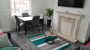Een woonkamer met een open haard, een eettafel met stoelen, een aan de muur bevestigde televisie en een raam met uitzicht op de straat.