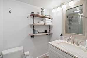 Et badeværelse med toilet, håndvask med spejl og to hylder med håndklæder og toiletartikler.