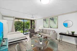 Et moderne hotelværelse med seng, spisebord i glas, sofa, skrivebord og balkon med udsigt over grønne områder.