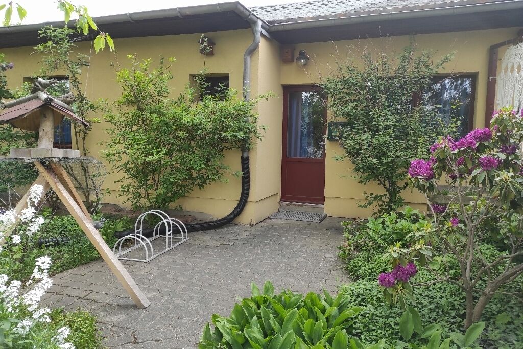 Ein gelbes Gebäude mit roter Tür, ein Garten mit verschiedenen Pflanzen und eine Metallkonstruktion.
