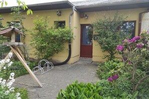 Ein gelbes Gebäude mit roter Tür, ein Garten mit verschiedenen Pflanzen und eine Metallkonstruktion.