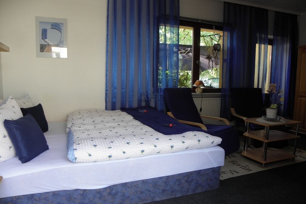 Ein Schlafzimmer mit einem Bett, einem Sessel, einem kleinen Tisch und einem Fenster mit Vorhängen.