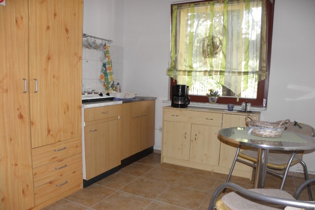 Eine Küche mit Holzschränken, einem kleinen Esstisch mit Stuhl, einem Fenster mit Vorhang und einer Kaffeemaschine.