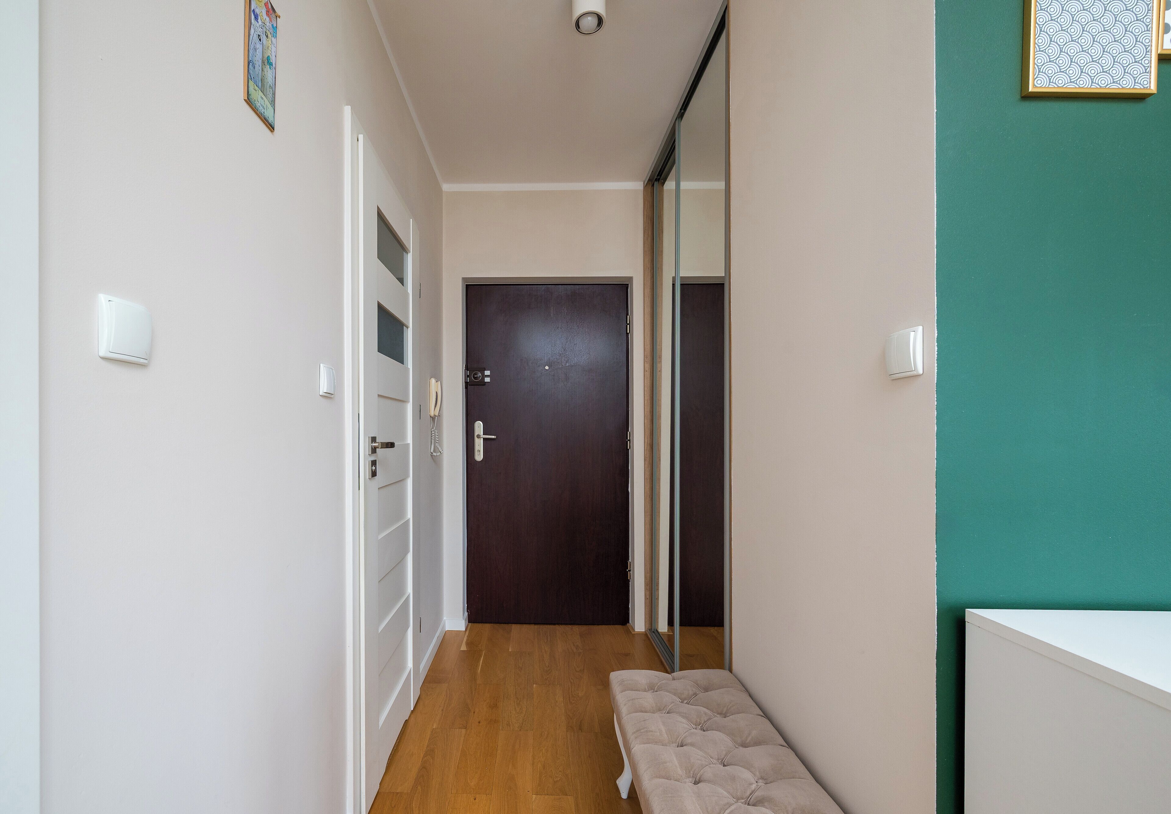 A hallway with a wooden floor, a bench, a door, and a mirror.