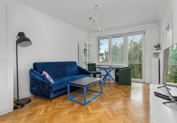 Ein modernes Wohnzimmer mit einem blauen Sofa, einem Couchtisch, einer Stehlampe, einem Esstisch mit Stühlen und einem Fernseher auf einem Schrank.