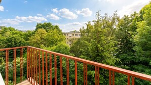 A balcony with a red railing overlooking a lush green landscape and a building in the distance. - Grzegórzki Cozy Apartment (Kraków)