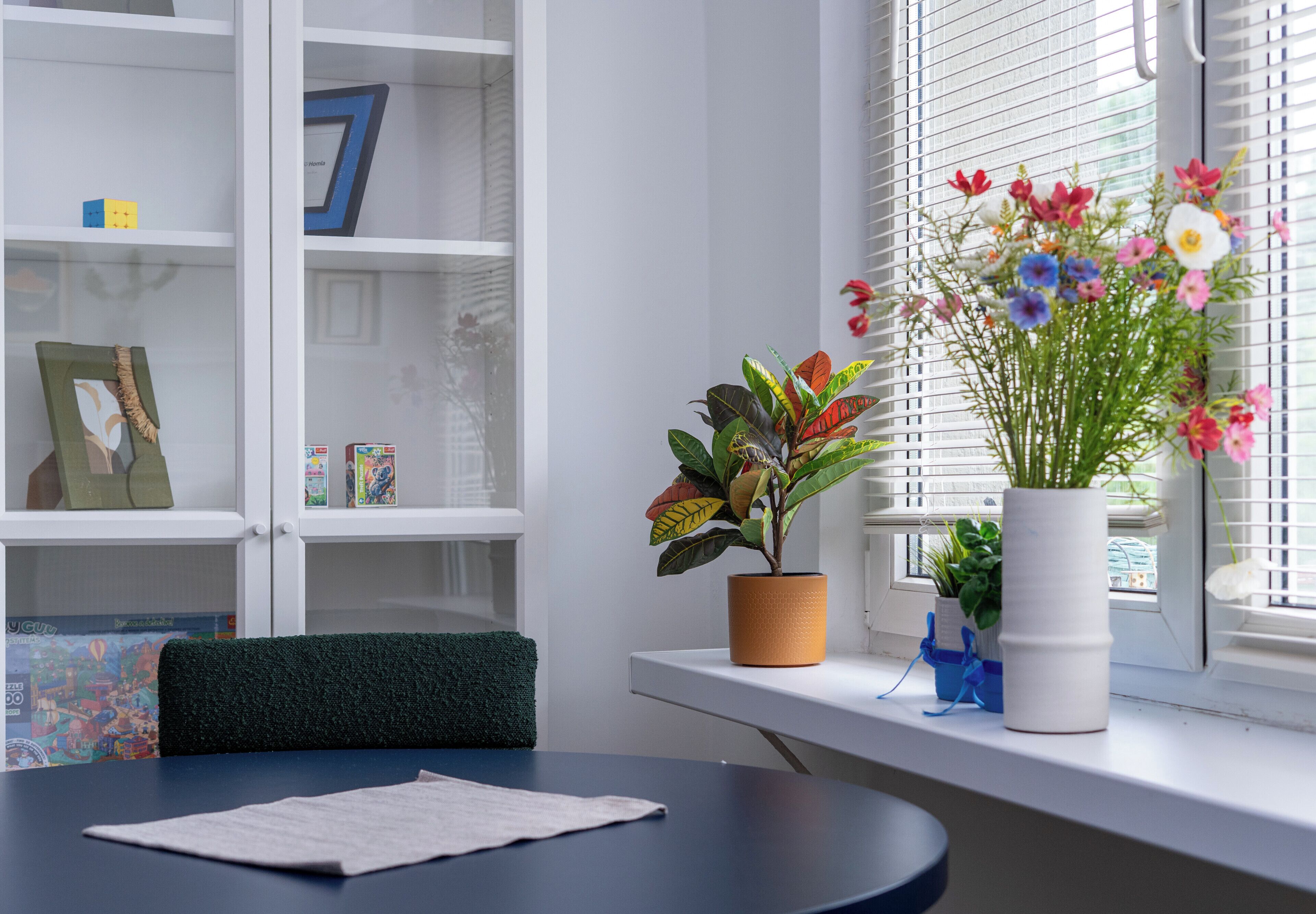 A room with a round table, a potted plant, and a window with blinds.