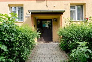 A building entrance with a paved pathway leading to a door, surrounded by dense green foliage. - Grzegórzki Cozy Apartment (Kraków)