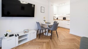 Ett modernt vardagsrum med en platt-TV, ett matbord med stolar, ett litet vitt skåp och ett köksområde i bakgrunden.