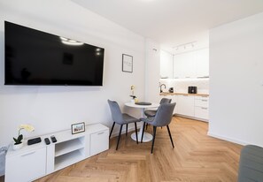 Ein modernes Wohnzimmer mit Flachbildfernseher, einem Esstisch mit Stühlen, einer kleinen weißen Kommode und einer Küchenzeile im Hintergrund.