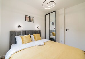 Una cama bien hecha con cabecera gris, ropa de cama blanca y amarilla, y un espejo en la pared.
