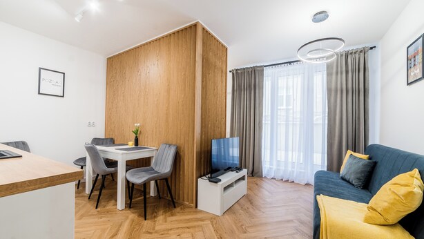 Een moderne woonkamer met een houten scheidingswand, een eethoek, een bank met een gele plaid en een televisie op een standaard.