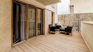 Een balkon met houten vloer, schuiframen en twee zwarte rieten stoelen.