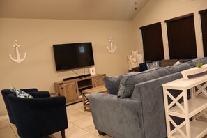 En stue med et fladskærms-tv, en grå sofa, en blå lænestol og et hvidt konsolbord.