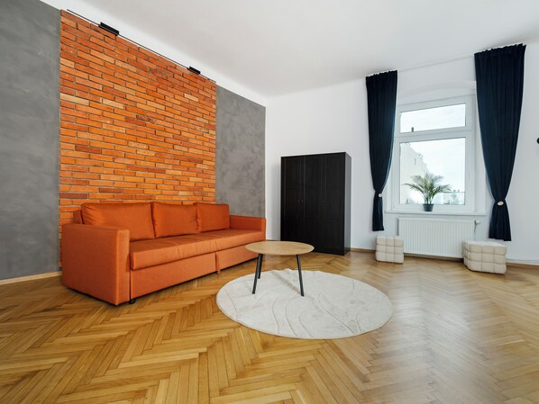 A modern living room with a herringbone wooden floor, a brick accent wall, a dark-colored wardrobe, and a window with curtains. - Łódź Homely Apartment (Łódź)