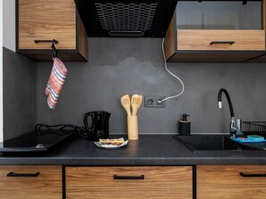 Eine Küche mit schwarzer Arbeitsplatte, Holzschränken, einer Kaffeemaschine, einem Wasserkocher, Holzkochlöffeln, einem schwarzen Wasserhahn und einem gestreiften Topflappen.