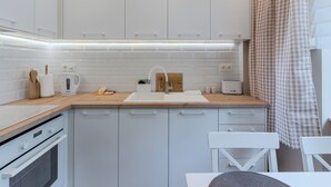 Een kleine keuken met witte kasten, een houten aanrecht, een inbouwoven en een eethoekje met een stoel.