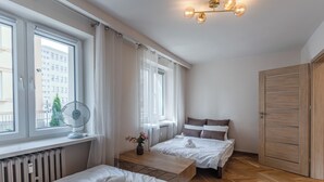 Een hotelkamer met twee bedden, een radiator, een ventilator, een houten bureau, een nachtkastje, een raam met gordijnen, een houten kledingkast en een plafondlamp.