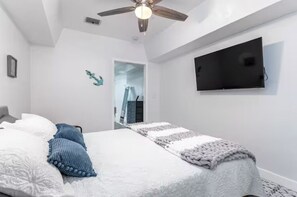 Un dormitorio con una cama, ventilador de techo, televisor y una puerta que da a otra habitación.