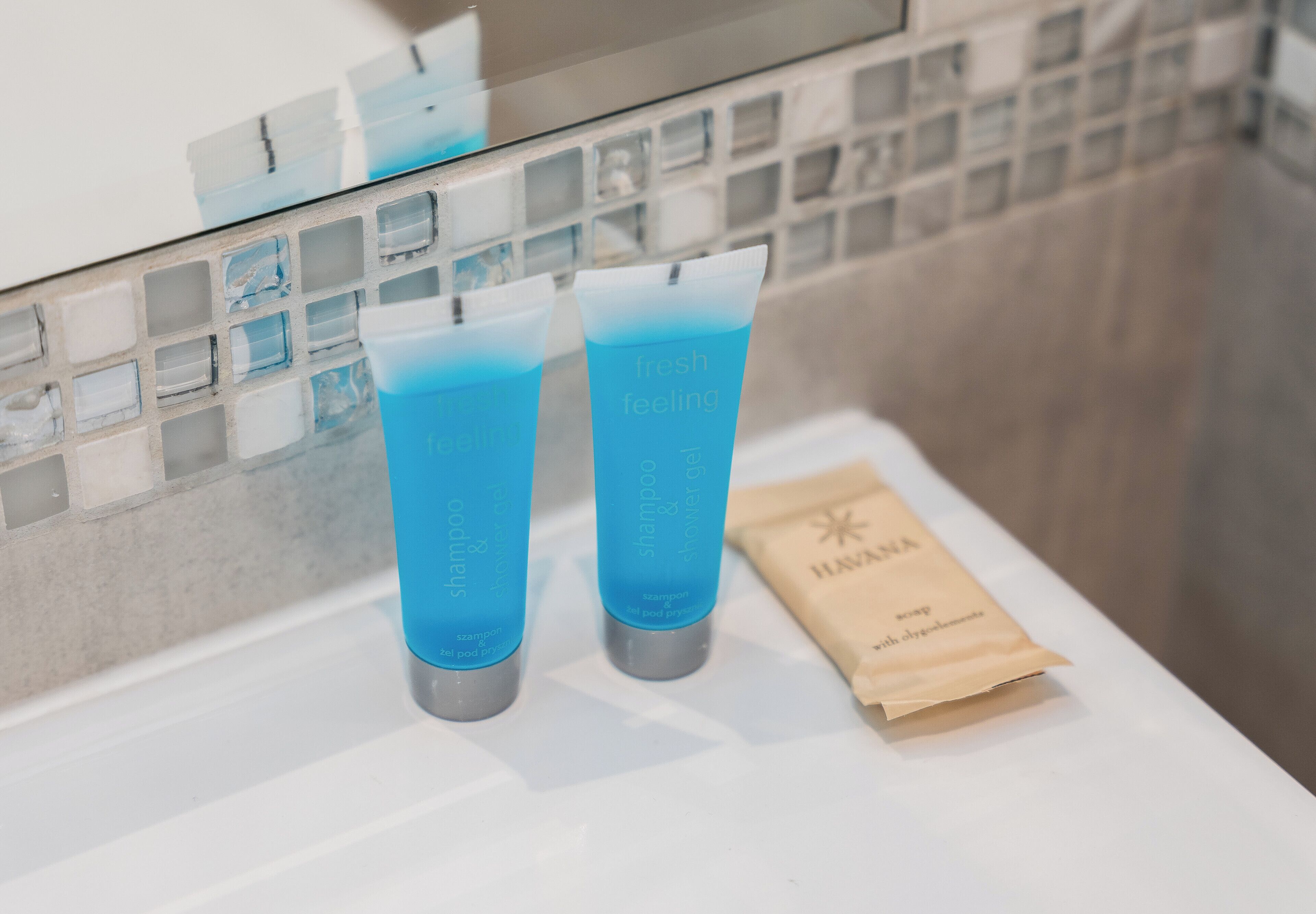 Deux tubes de shampoing et d’après-shampoing bleus sont posés sur une surface blanche, à côté d’un sac en papier marron.