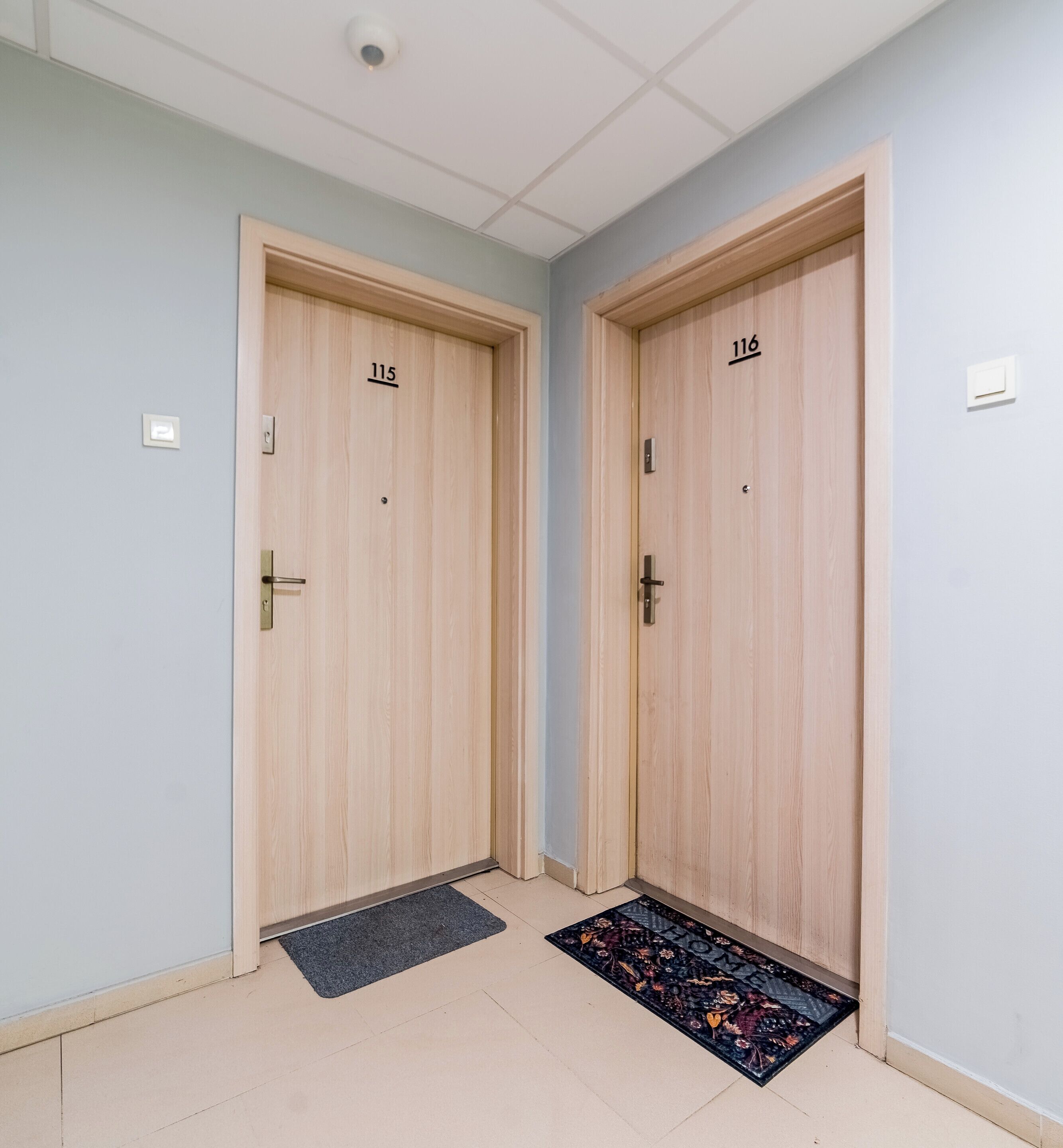 Two wooden doors with numbers 115 and 116, a doormat, and a light blue wall.