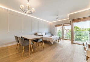 Une chambre moderne avec un lit, une table de chevet et une vue sur la verdure grâce à de grandes fenêtres.